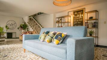 A living room with a blue sofa, patterned cushions, a staircase, a bookshelf with hats, and a wall with a sign.