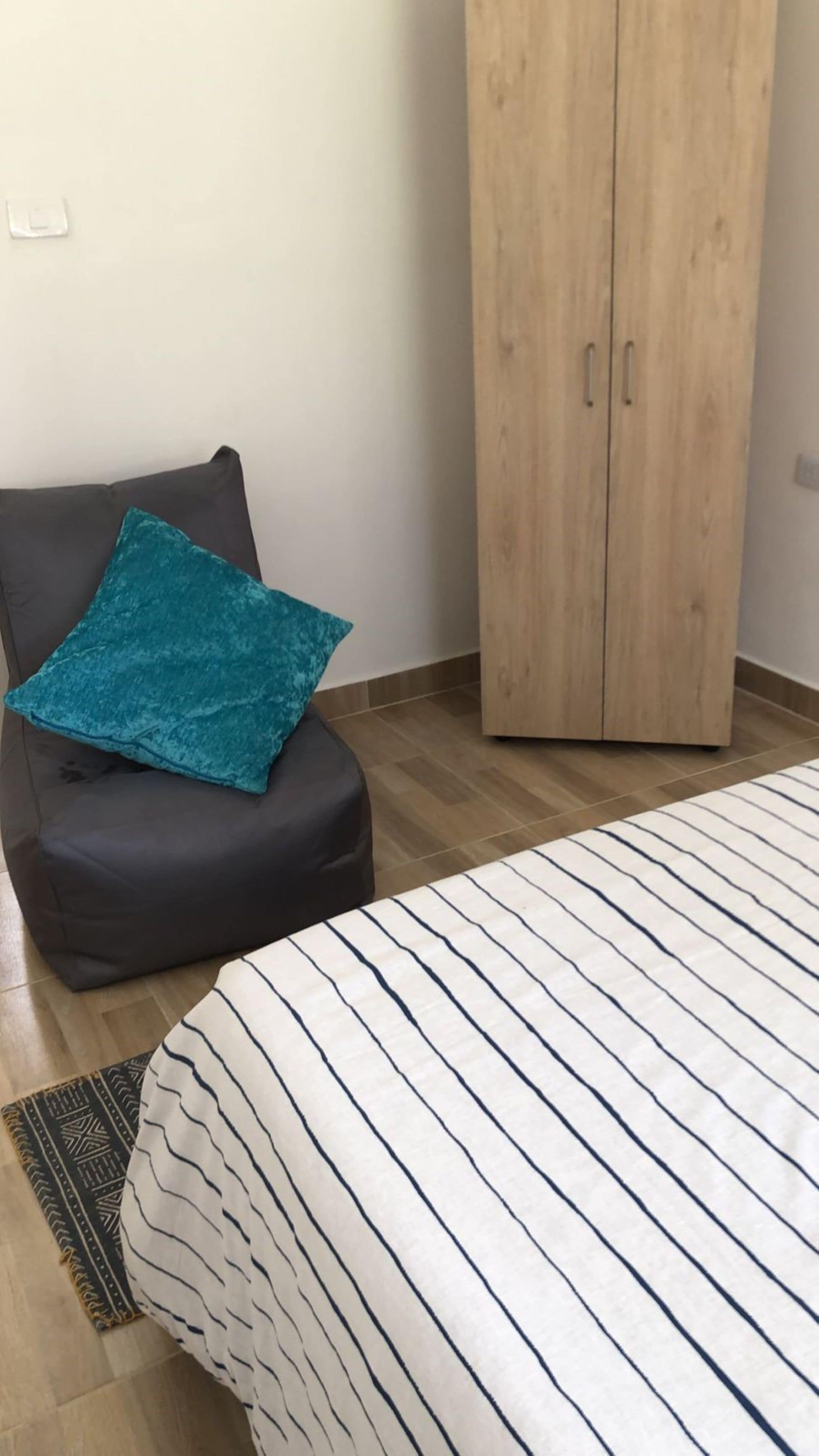 Un dormitorio con cama, una silla negra con cojín verde azulado, un armario de madera y una alfombra estampada.
