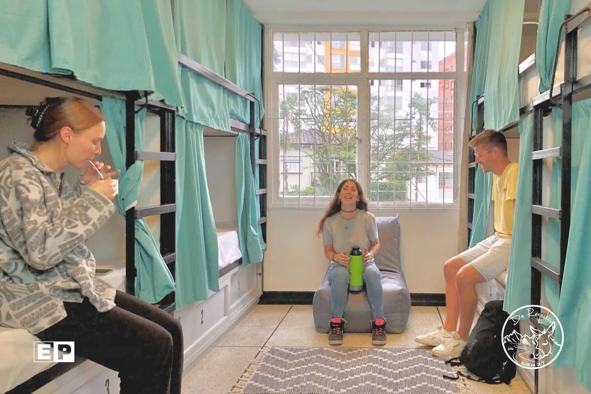 A dormitory room with bunk beds, a window with curtains, and a patterned rug on the floor.