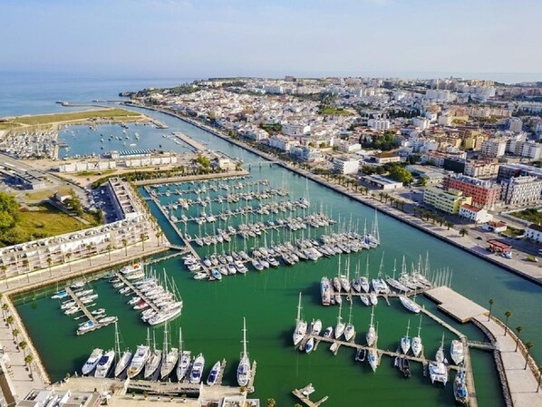 An aerial view of a marina with numerous boats docked, surrounded by urban buildings and a clear sky.