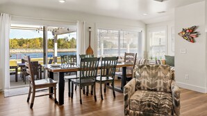 A dining area with a wooden table and chairs, a sofa, and a view of a sports field through large windows. - Always Channel-On Muskegon Channel, walk to lighthouse, beach, the DECK & swim.  (Muskegon)