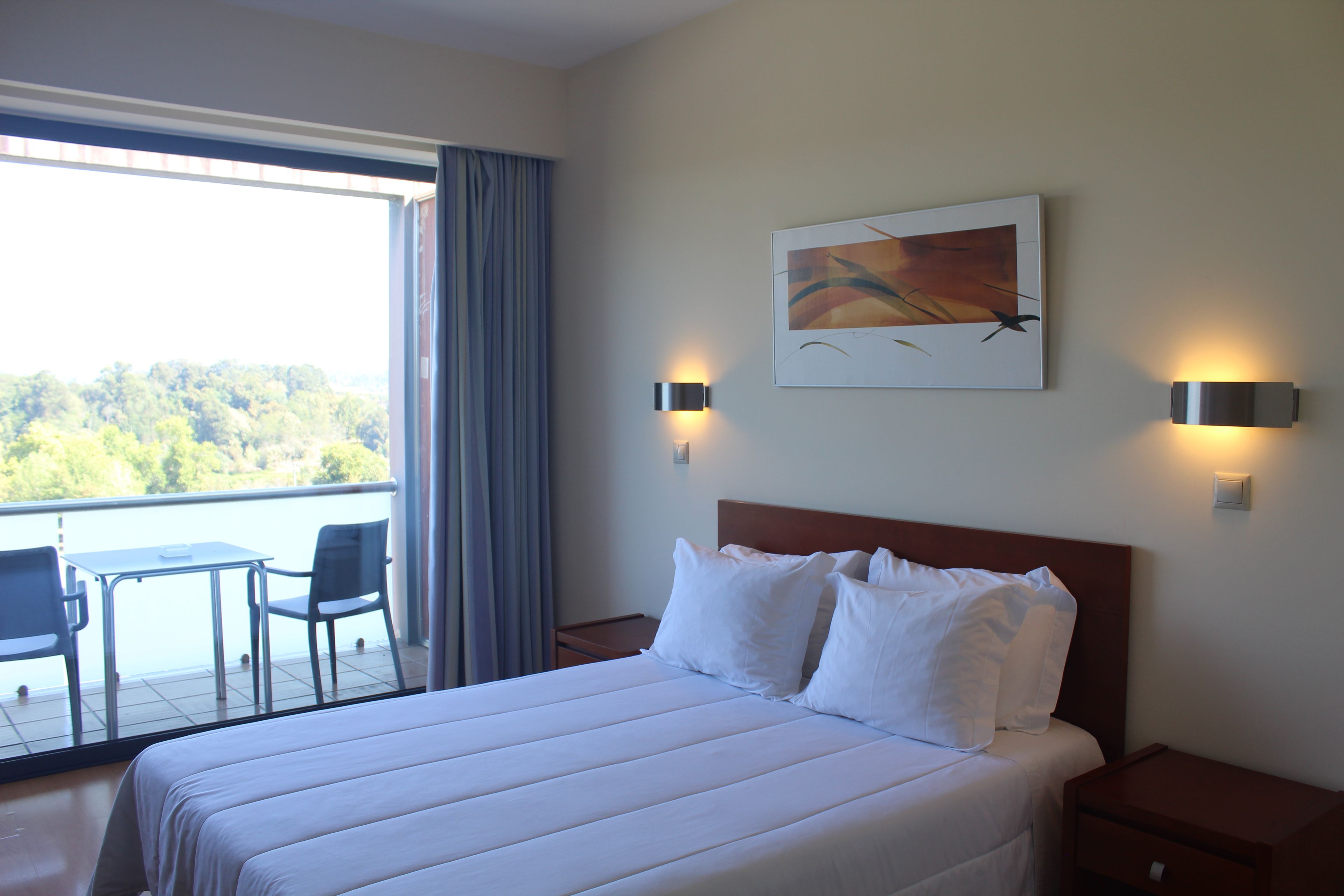 A hotel room with a bed, a desk, and a chair. There is a window with a view of trees and a balcony.