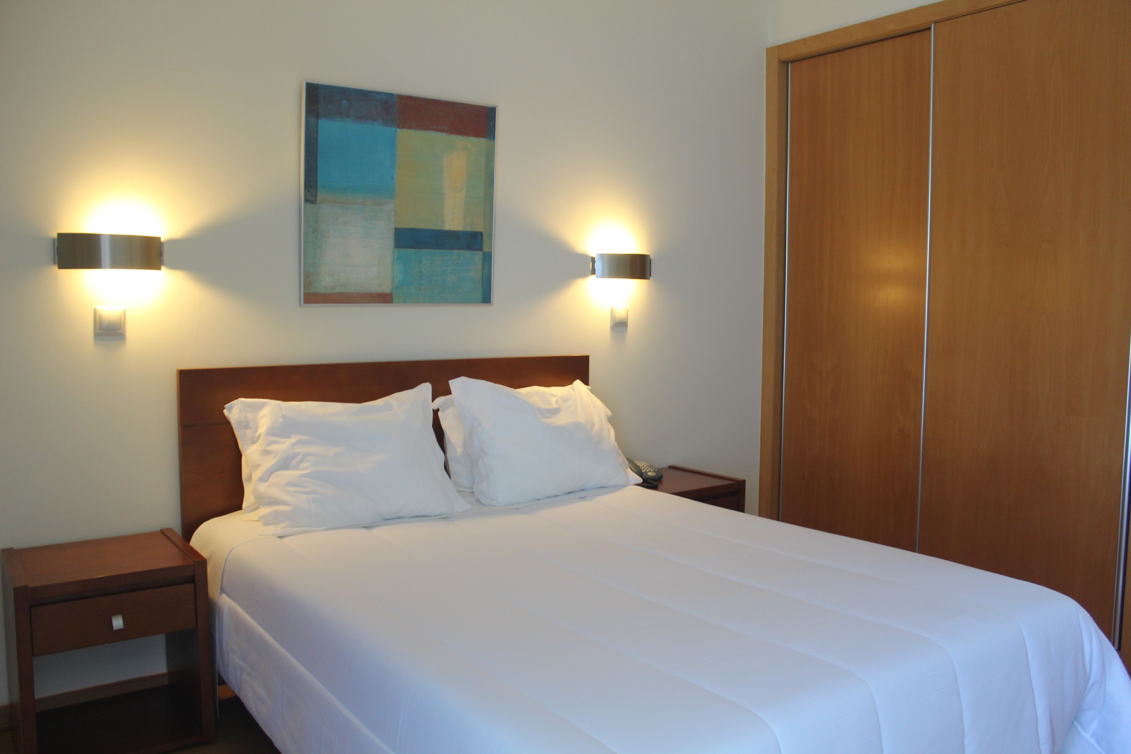 Habitación de hotel con cama, dos lámparas de noche, mesita de noche, armario de madera y un cuadro en la pared.