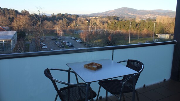A balcony with a table and two chairs, overlooking a parking lot and a hilly landscape. - Hotel Dom Afonso (Monção)