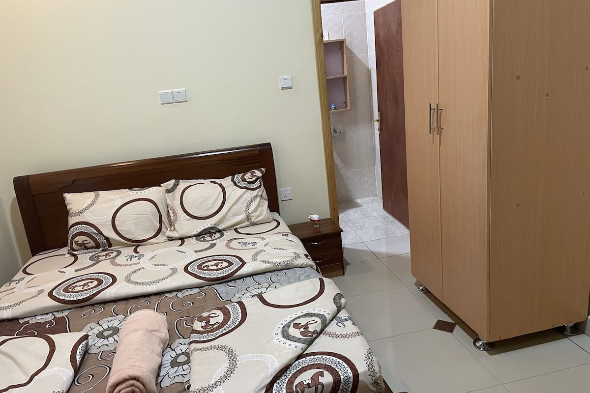 A neatly made bed with patterned bedding, a wooden wardrobe, and a tiled floor.