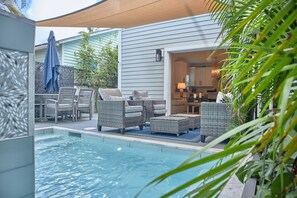 A poolside area with wicker furniture, a covered seating area, and a view into a house.