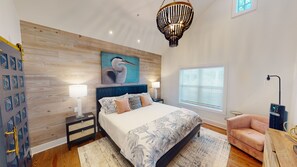 A bedroom with a wooden wall panel, a bed with a floral bedspread, a nightstand with a lamp, a painting of a bird, a window with blinds, and a pink chair.
