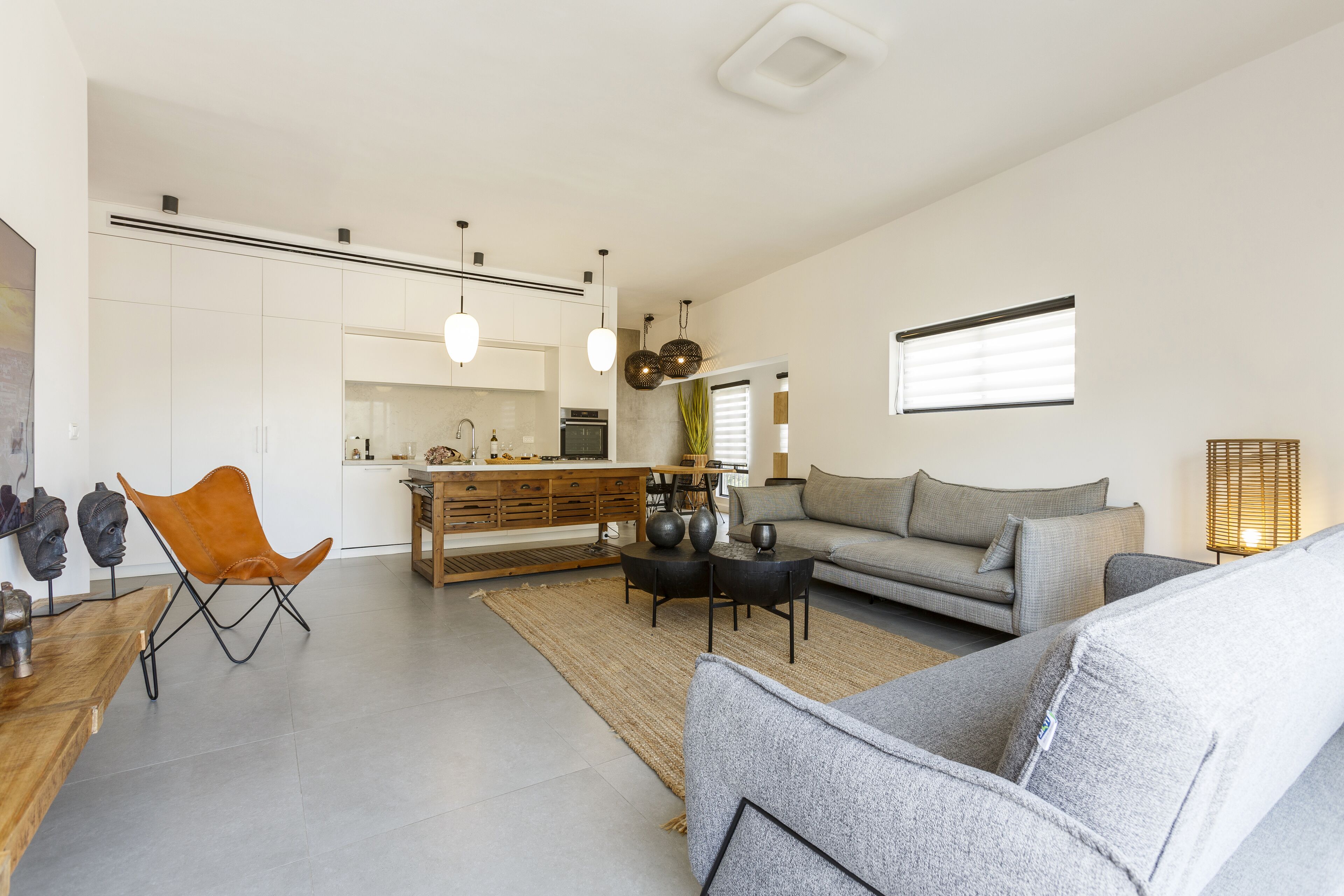 A modern living room with a grey sofa, a wooden coffee table, and a brown leather chair. There is a kitchen in the background with white cabinets and a dining area.