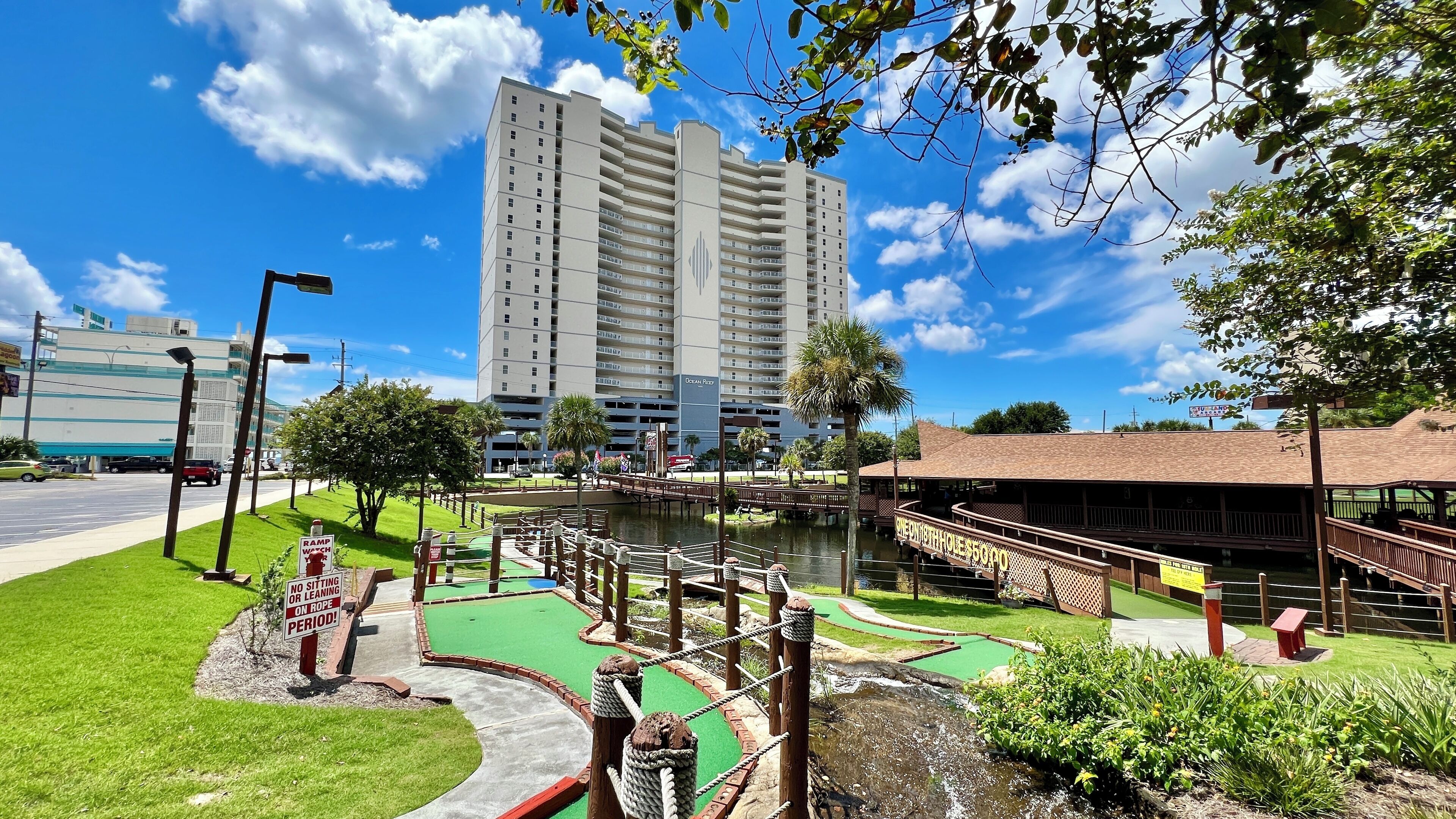 Een golfbaan met een waterhindernis, een houten brug en een gebouw met de naam 'Ocean Reef' op de achtergrond.
