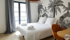 Une chambre d’hôtel comprenant un lit, une chaise, une table à manger, une grande fenêtre avec des rideaux et une fresque murale représentant des palmiers.