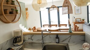 Une cuisine moderne avec un plan de travail en marbre, une machine à café et un évier. On y trouve des étagères en osier ornées de plantes et deux lampes suspendues en osier.