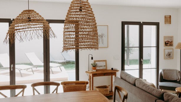 Un espace repas comprenant une table et des chaises en bois, un canapé et deux suspensions lumineuses en fibres tressées.