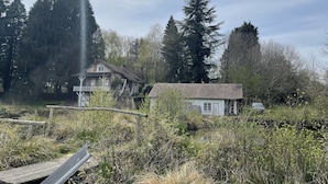 Ett rustikt hus omgivet av tät skog, med en liten damm i förgrunden och en träbro som leder fram till huset.