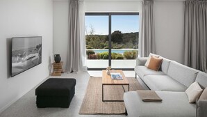 A modern living room with a grey sofa, black ottoman, and a coffee table. A large window offers a view of a pool and greenery. - Villa Sealine in Vinkuran (Vinkuran)