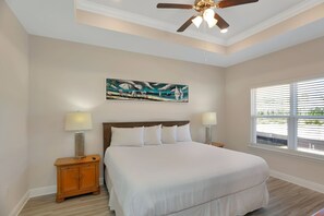 A bedroom with a large bed, two bedside tables with lamps, a ceiling fan, a painting on the wall, and a window with blinds.