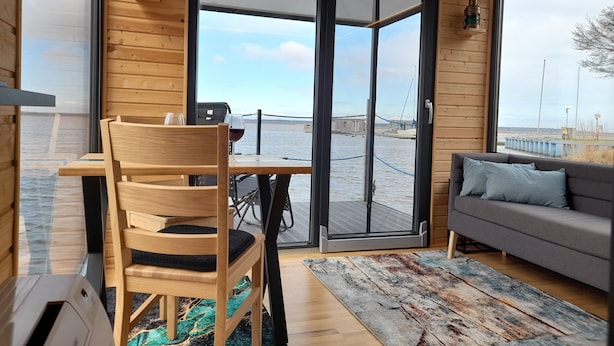 A modern interior with a wooden wall, a sofa, a dining table with chairs, and a view of the water outside.