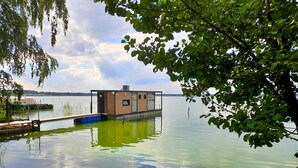 A floating house with a wooden exterior, docked on calm water, surrounded by trees and a clear sky.