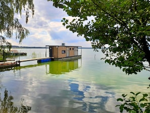 Et flydende hus med træfacade, fortøjet i rolige vand, omgivet af træer og en klar himmel.