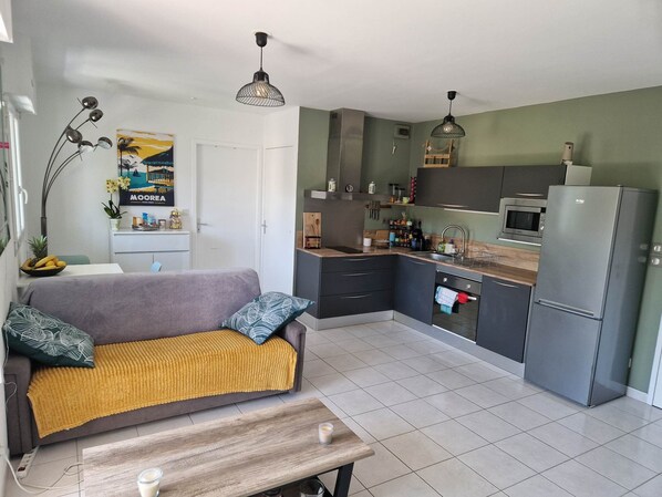 A modern kitchen with grey cabinets, a microwave, and a refrigerator. There is a wooden dining table with a bench, a sofa with a mustard-colored throw, and a decorative plant.