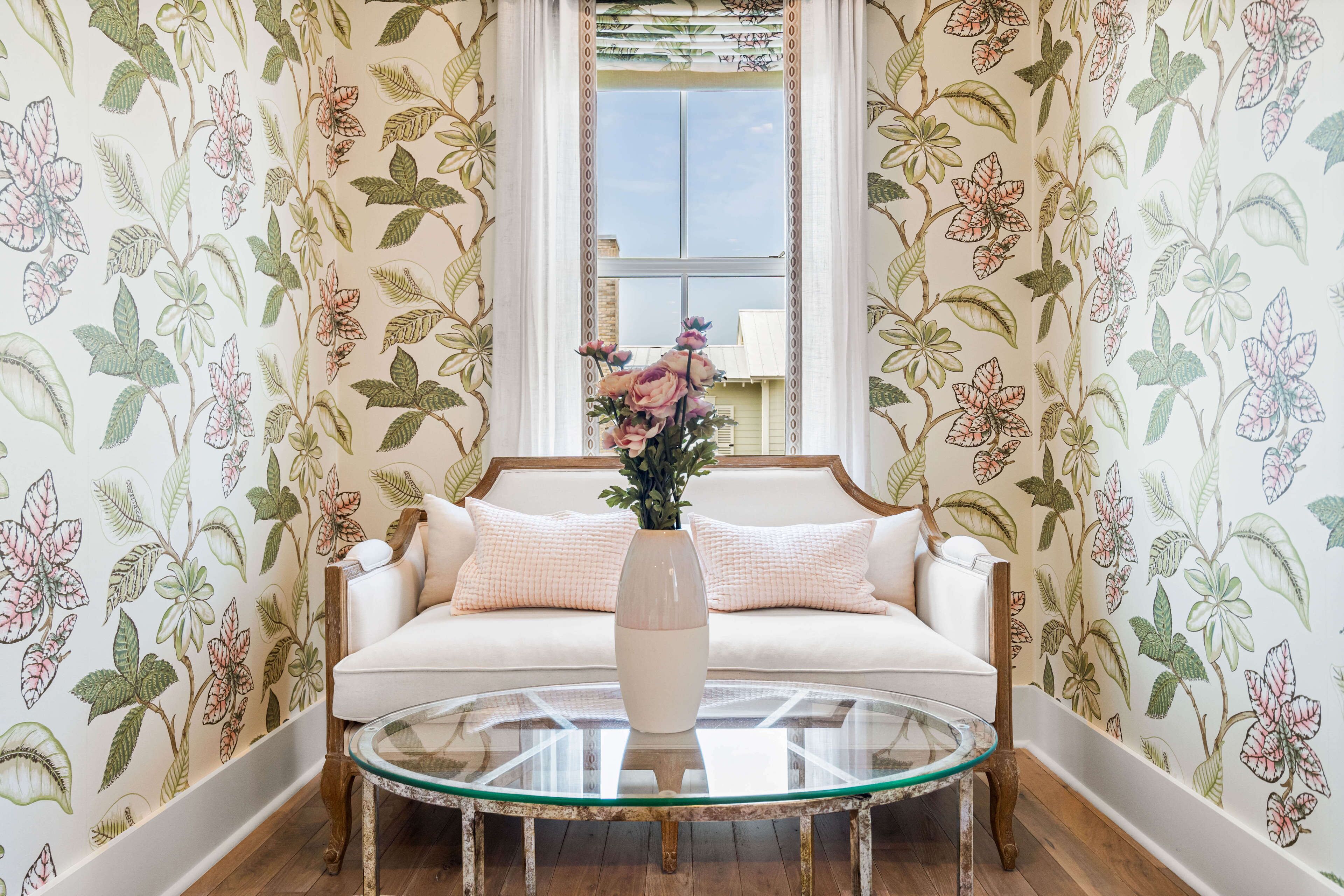 A room with floral wallpaper, a white sofa with pink cushions, and a glass coffee table.