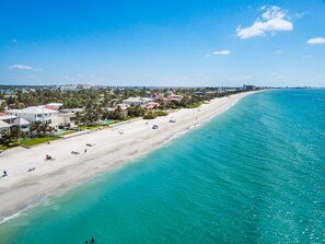 A coastal beach area with a sandy shore, clear turquoise waters, and a row of buildings along the beach.