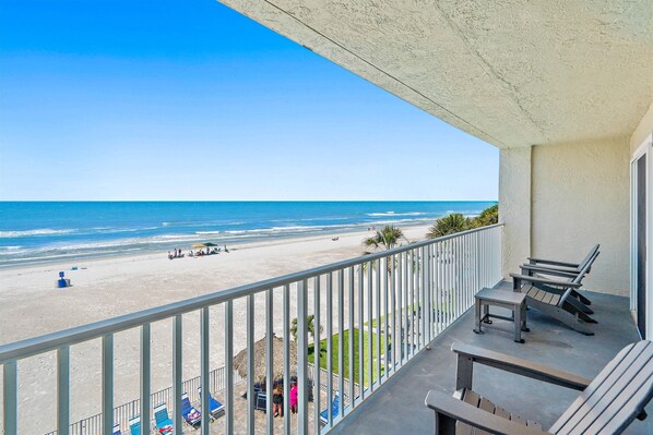 A balcony with a view of the beach, lounge chairs, and a table.