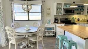 A kitchen with a round table, white chairs, a coffee maker, a microwave, and a wooden island with stools. - HeartWind Cottage -Quaint and cozy with private pool, steps to the beach (Santa Rosa Beach)