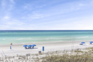 A beach with clear skies, calm waters, and a sandy shore. There are blue umbrellas and beach chairs set up for relaxation.