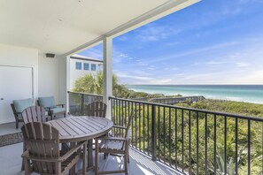 Un balcón con mesa y sillas de madera, con vista a una playa y una calzada que conduce al agua.