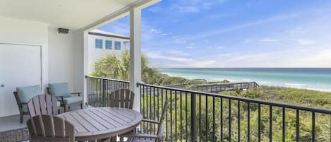 A balcony with a wooden table and chairs, overlooking a beach with a clear view of the ocean and a pathway leading to the water.