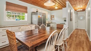 A modern kitchen with a wooden ceiling, white cabinets, a large wooden dining table with chairs, and a window with blinds.