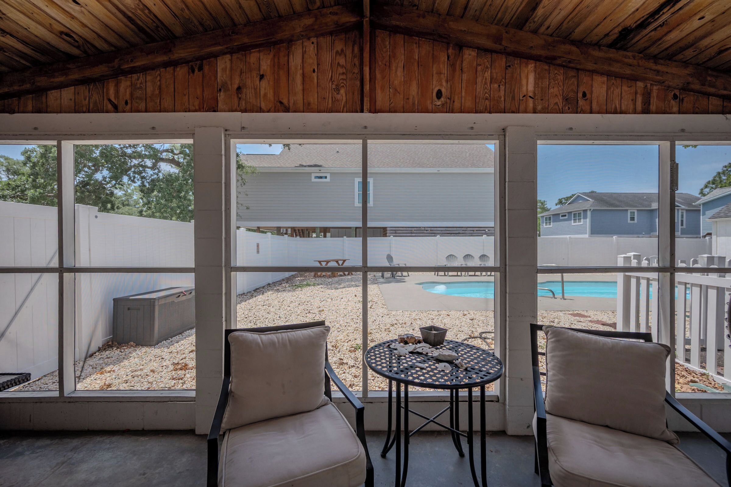 A covered patio with two chairs and a table, a pool in the background, and a view of neighboring houses.