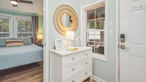 A bedroom with a bed, a white dresser with drawers, a round mirror, a window with curtains, and a door with a lock.