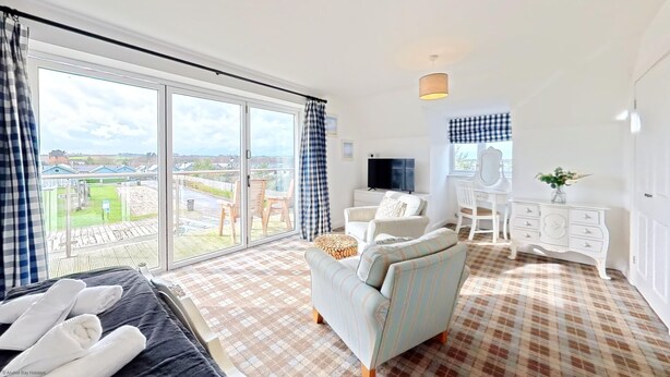 A spacious room with a large sliding glass door leading to a balcony, a bed with white towels, a light blue armchair, a white dresser, and a television.