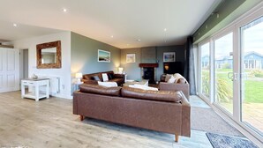 A spacious living room with a large brown sofa, a fireplace, and a large window offering a view of the outside.