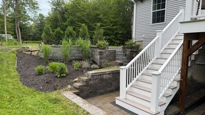 Una casa con un muro de piedra, un jardín con diversas plantas y una escalera blanca que conduce a la entrada.