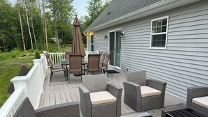 Un patio exterior techado con muebles de mimbre y una mesa, junto a una casa con terraza y barandilla blanca.