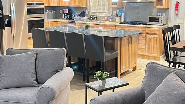 Cocina moderna con armarios de madera, un refrigerador de acero inoxidable y una isla central con asientos. Hay dos sofás grises y una pequeña mesa con una planta.