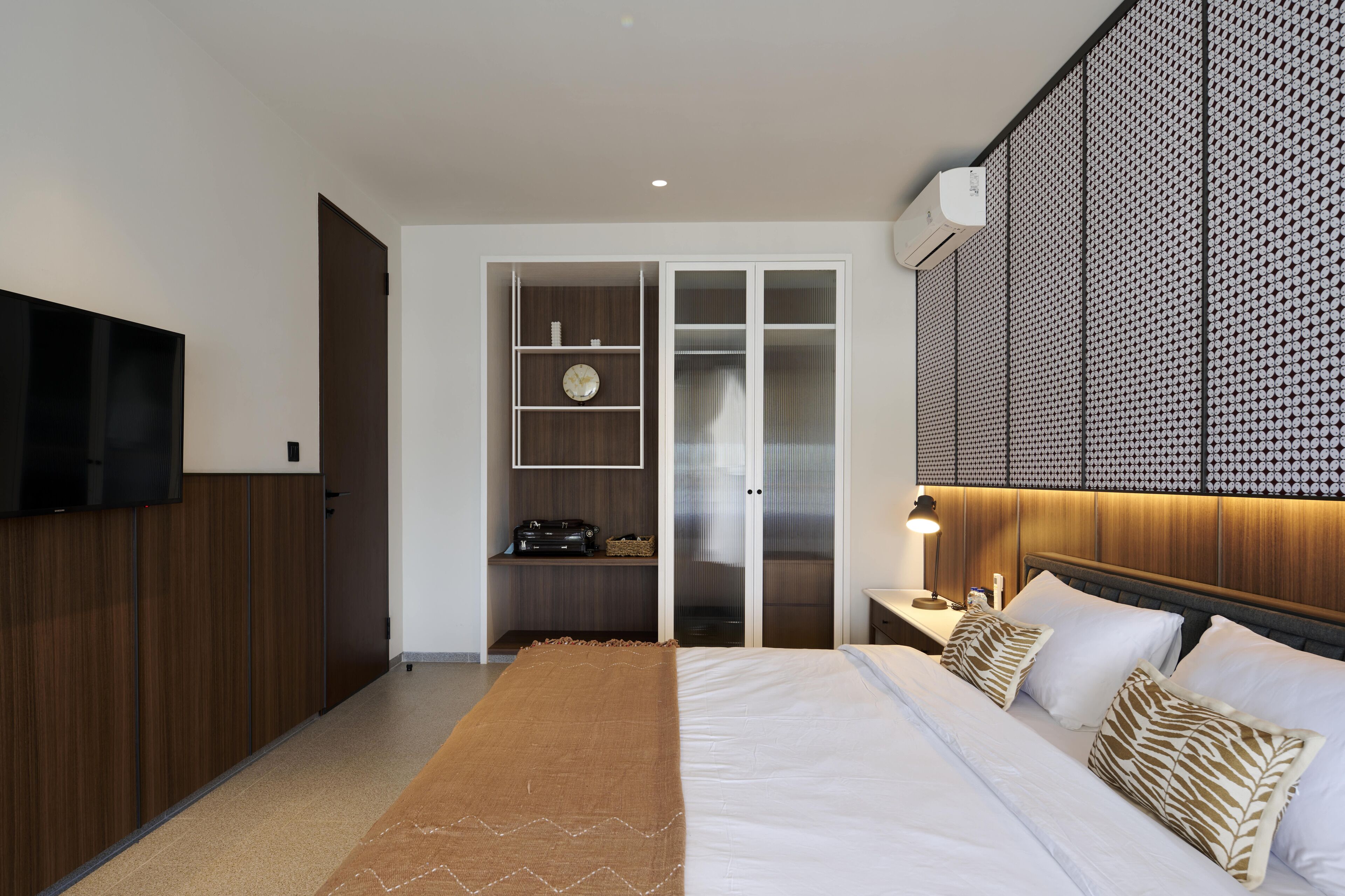 Une chambre moderne avec un grand lit, un téléviseur fixé au mur, une armoire et un petit bureau avec une lampe.