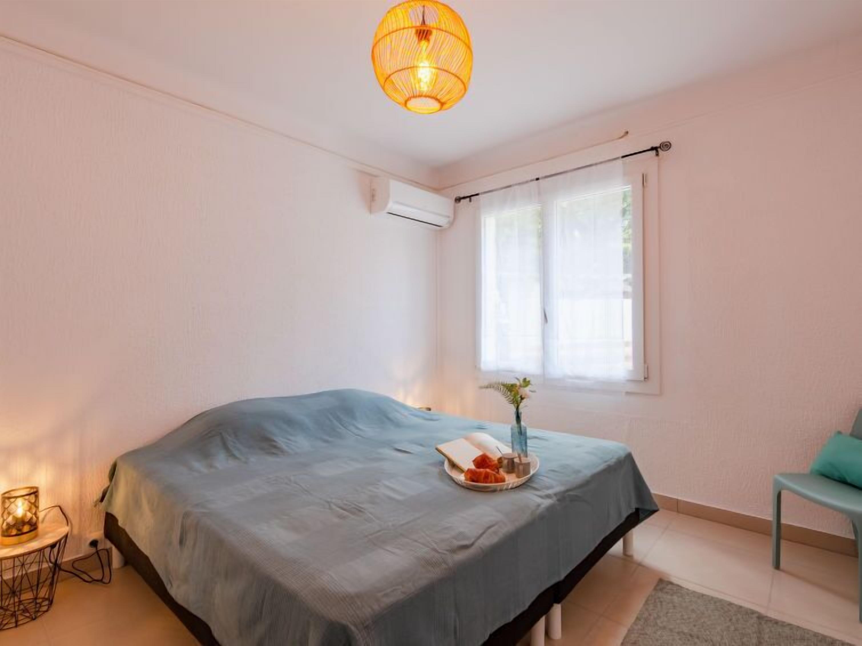 Un dormitorio con cama, mesita de noche con lámpara, una silla y ventana con cortinas.