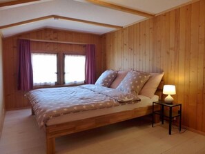 Quarto com painéis de madeira, uma cama, duas janelas com cortinas e uma mesa de cabeceira com abajur.