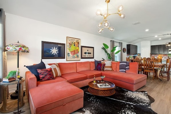 A modern living room with a sectional sofa, a coffee table, and a chandelier. There are framed pictures on the wall and a plant in the corner.