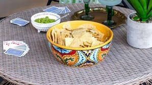 Una silla de mimbre con cojines, una mesita con un cuenco de papas fritas, una planta en maceta y una ventana con vista a un edificio.