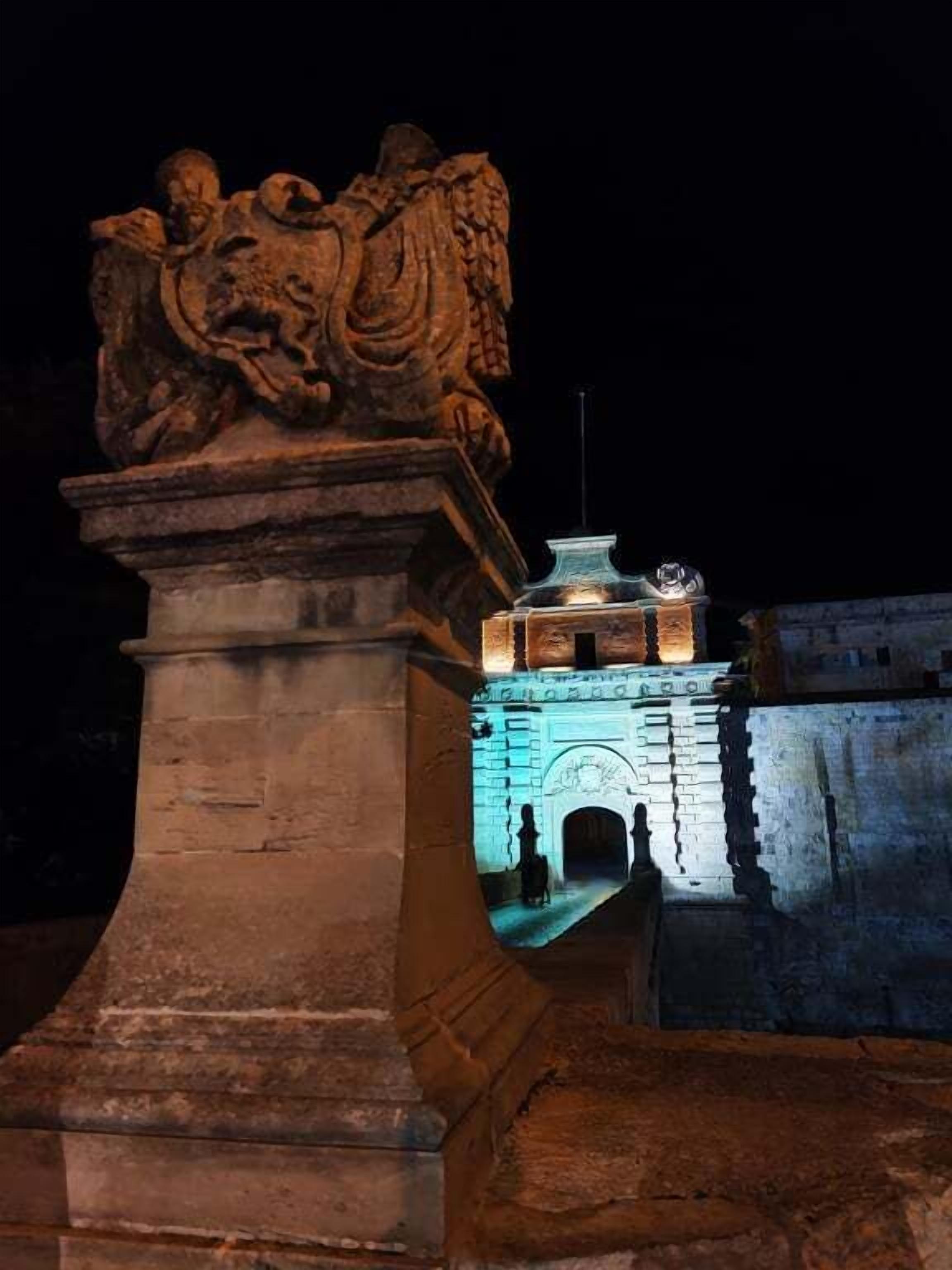 Un monument en pierre orné d’une sculpture en relief, une petite structure illuminée surmontée d’un mât pour drapeau, et un mur en pierre doté d’une entrée en arc de cercle.