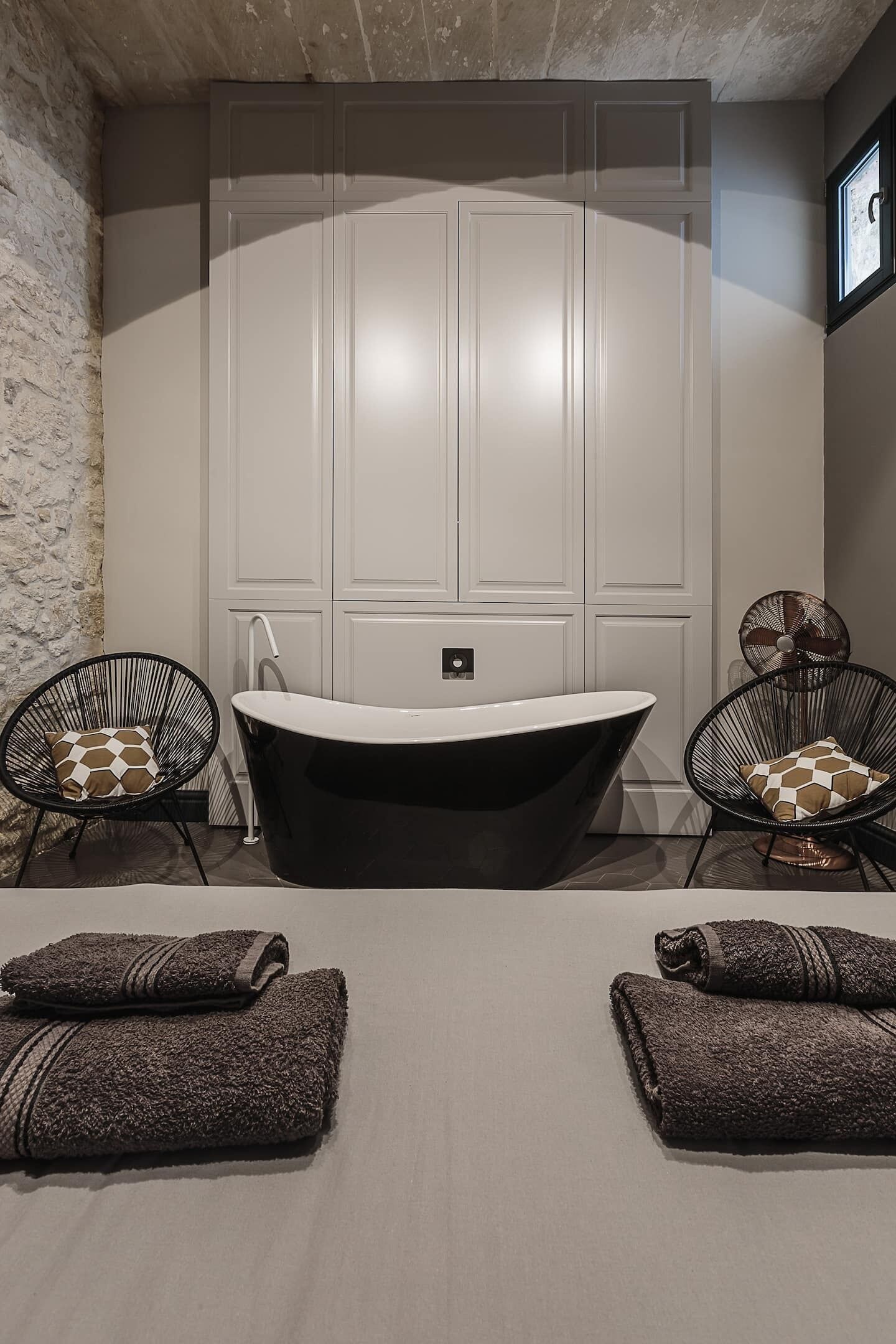 Une salle de bain avec une baignoire sur pieds, deux chaises noires en fil de fer avec des coussins à motifs, et deux serviettes enroulées posées sur le sol.