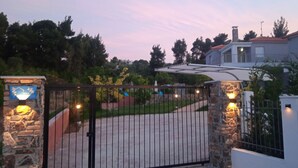 Una entrada cerrada con pilares de piedra y un letrero azul, rodeada de follaje y una casa con techo rojo.