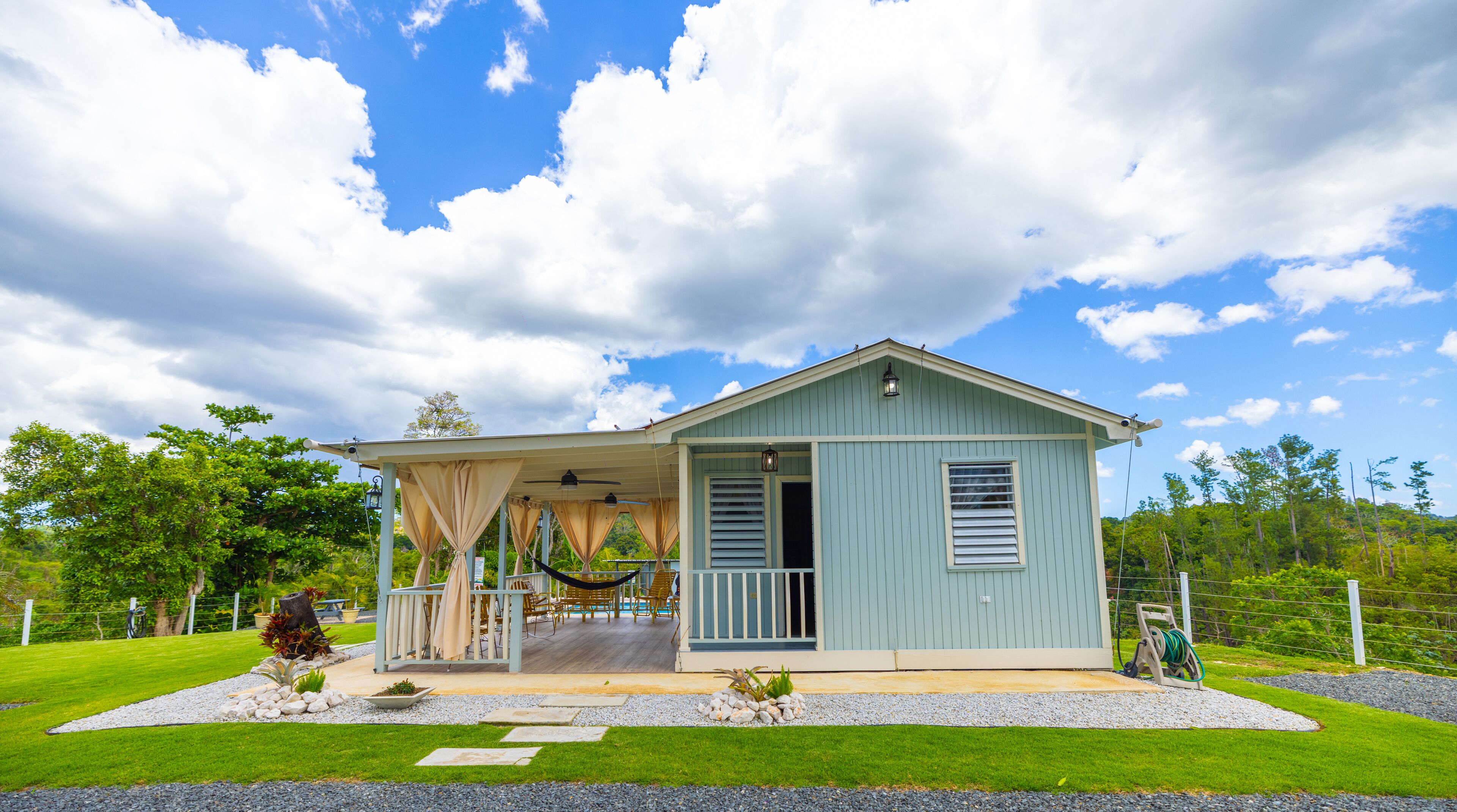 A light blue single-story house with a covered porch, a small deck, and a garden with a lawn and gravel paths.