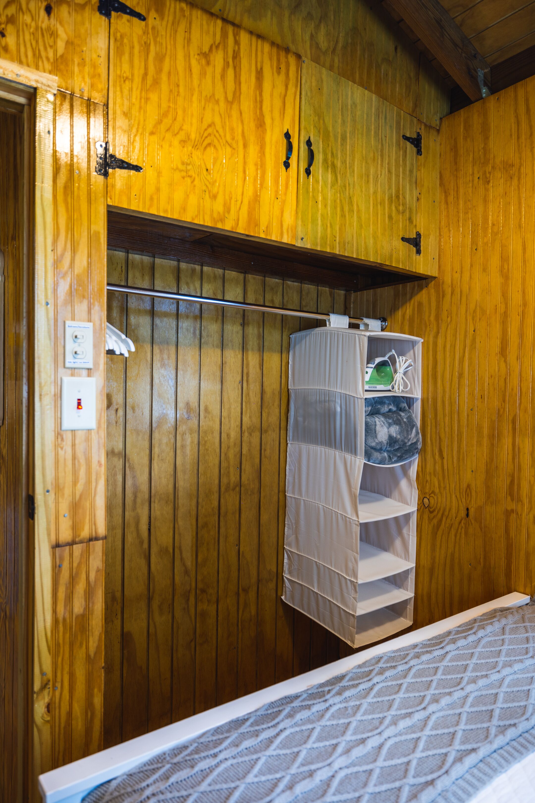 A wooden-paneled room with a bed, a hanging storage unit, and a visible electrical outlet.