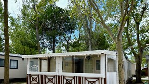 Una casa móvil de una sola planta, pequeña, con fachada blanca y detalles en marrón, rodeada de árboles.