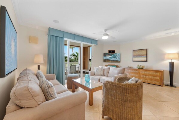 A living room with a sofa, coffee table, and wicker chair. There is a TV, a painting, and a lamp.